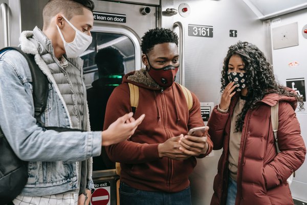 There are three young adults with their phone standing in the train with their masks on.